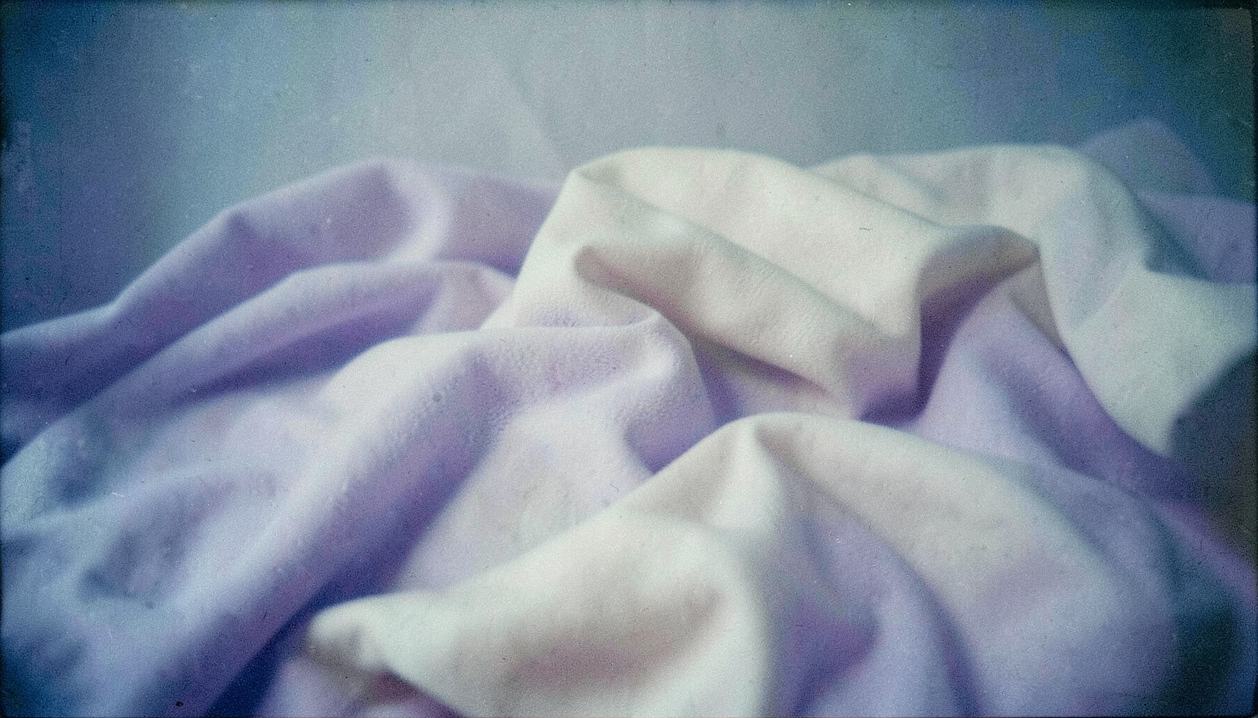 A calm, muted-toned still life with soft lavender and cream fabric folds, suggesting rest and self-care.